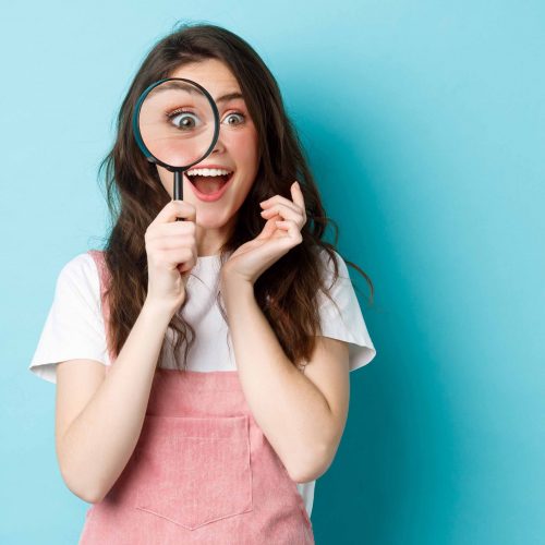 Happy young woman looking through magnifying glass with excited face, found or search something, standing over blue background Happy young woman looking through magnifying glass with excited face, found or search something, standing over blue background.